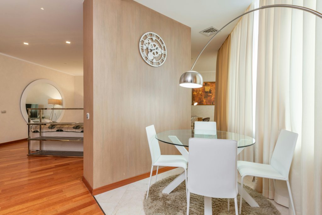 Pourquoi la visite virtuelle 360° aide à vendre votre bien plus rapidement Dining zone with glass round table and white chairs separated from living room with wooden wall decorated with creative clock in daylight